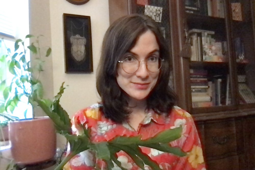 Annette smiles with a houseplant in her lap as she sits in front of a bookcase and a window
